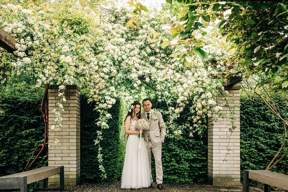 A bride and groom stand together outdoors beneath a floral arch made of cascading white blossoms and green foliage. The bride is holding a bouquet of soft pink and white flowers, dressed in a white we