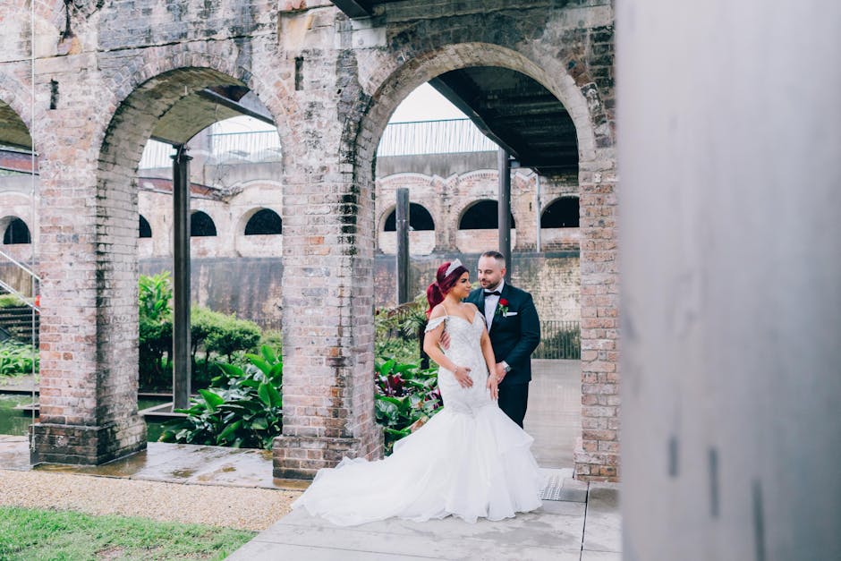 A bride and groom stand beneath a series of brick arches in an outdoor setting, with lush green plants and water features visible in the background. The bride wears a white, off-the-shoulder wedding g