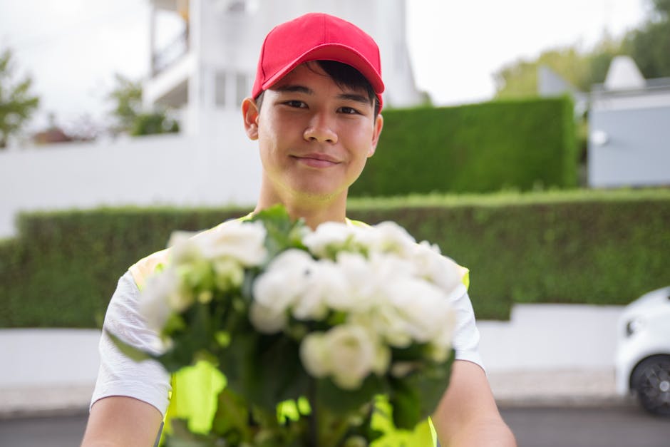 A smiling delivery person wearing a red cap and a high-visibility vest holds out a lush bouquet of predominantly white flowers, including roses and lisianthus, wrapped in clear and green paper. The ar