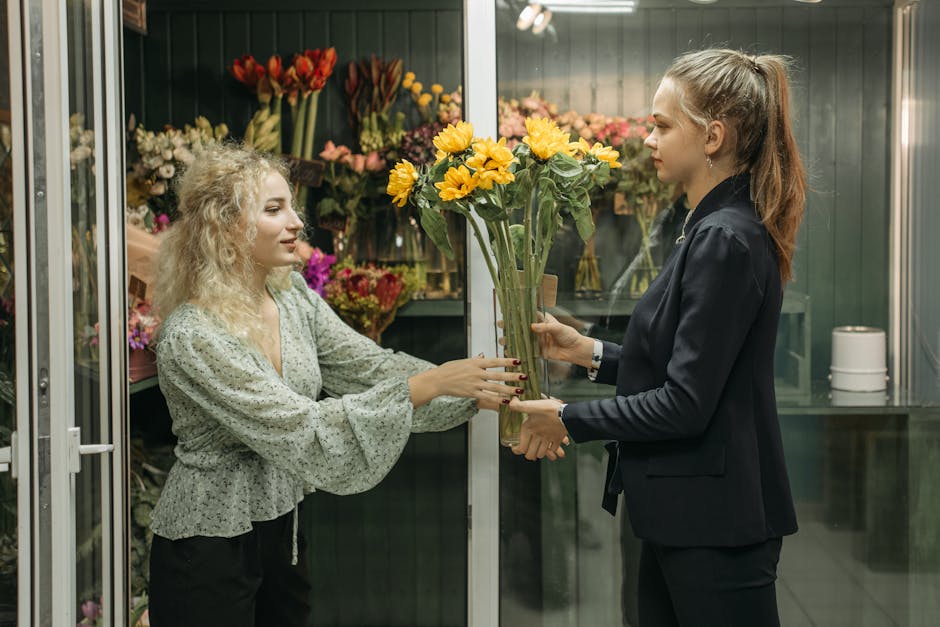 A woman with curly blonde hair wearing a light-colored, floral blouse is inside a flower shop, selecting a bouquet of vibrant yellow sunflowers arranged in clear glass vases. She is reaching out to ac