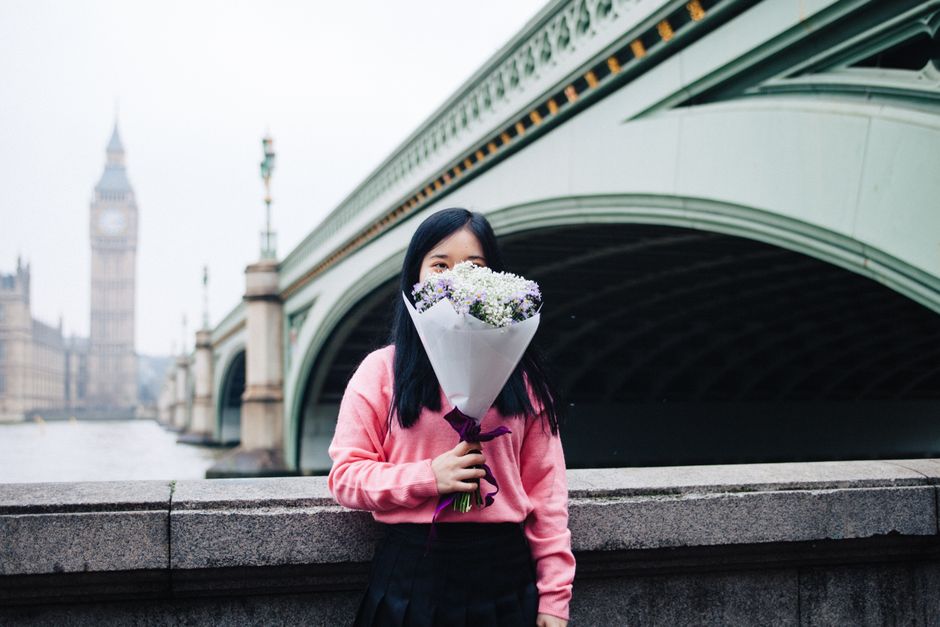 A woman with long black hair wearing a pink sweater and a black skirt is holding a bouquet of fresh white and purple flowers wrapped in white paper and tied with a dark purple ribbon. She stands near