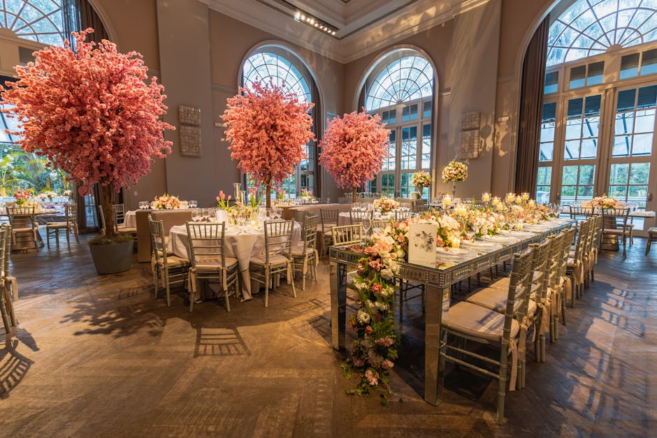 A spacious, elegantly decorated venue with large arched windows allowing natural light to illuminate the room. The interior features a mix of round and rectangular tables covered with white tablecloth