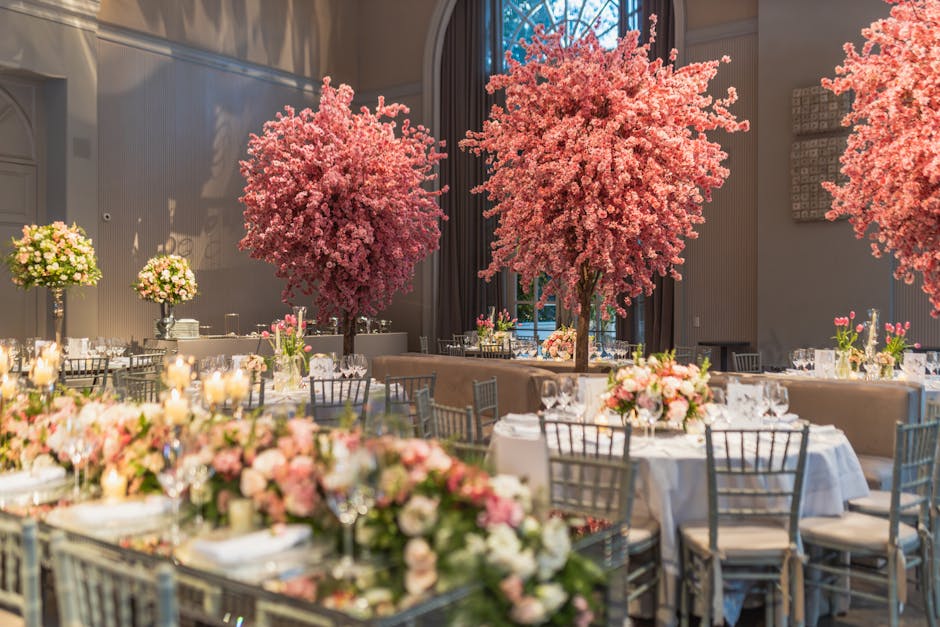 An elegant indoor event space decorated with lush pink blossoming trees and floral arrangements. The room features round tables draped with white tablecloths, each adorned with pastel-colored roses, p
